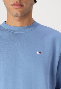 Sudadera azul claro con un acabado texturizado, cuello de canalé y pequeño logo bordado en rojo, blanco y azul en el pecho.