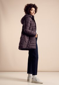 Puffjacka i mörklila med huva, med horisontell quiltning. I kombination med mörka jeans och vita sneakers, mot en neutral bakgrund.