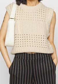 Krémový pletený top bez rukávů s texturovaným vzorem, žebrovaným výstřihem a volným střihem, spárovaný s černými kalhotami s proužky.