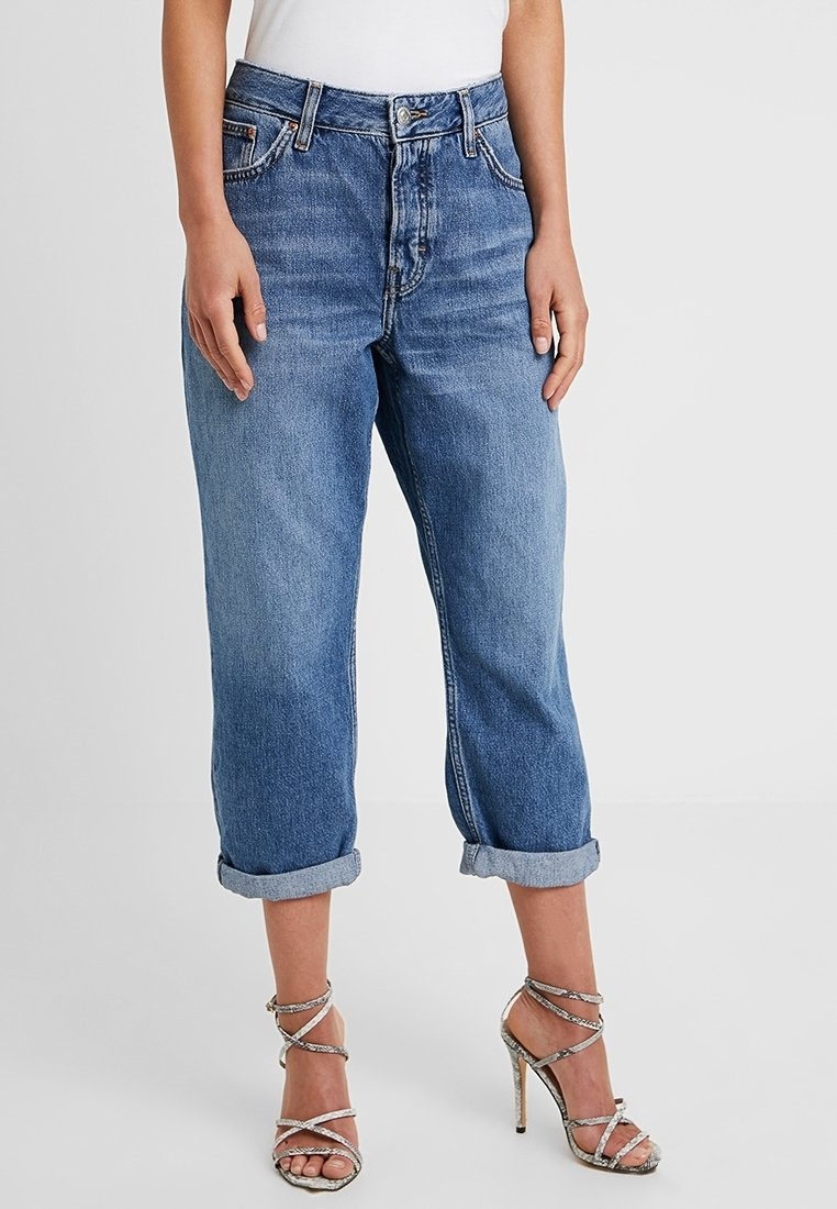 Světle modré, opotřebované džíny s volným střihem, srolovanými nohavicemi, předními kapsami a knoflíkovým zapínáním. Spárovány s stříbrnými sandály na vysokém podpatku.