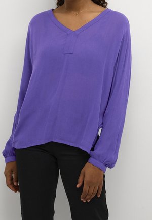 Personne portant un chemisier violet ample à manches longues avec un col en V et un pantalon noir sur un fond clair uni.