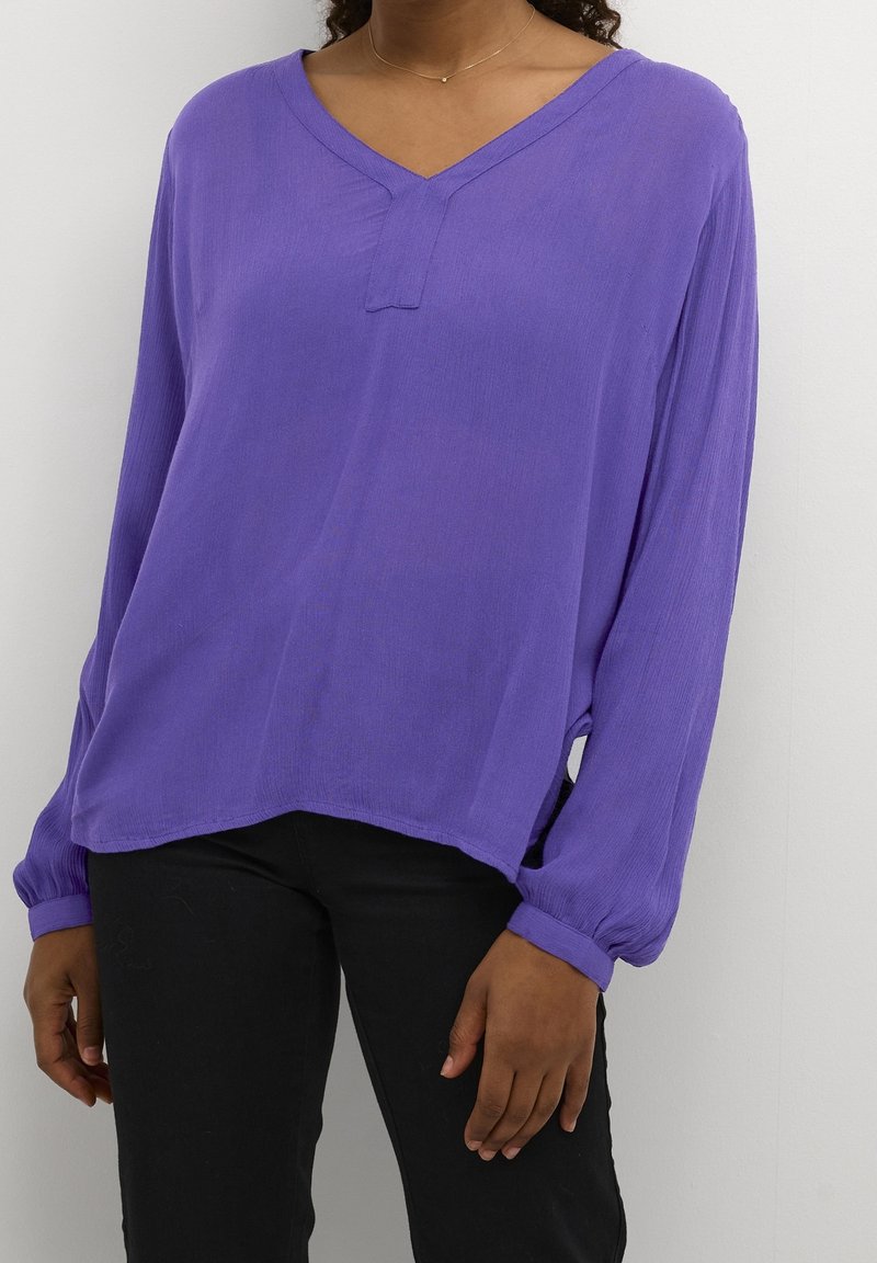 Personne portant un chemisier violet ample à manches longues avec un col en V et un pantalon noir sur un fond clair uni.