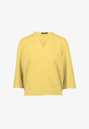 Gul kortærmet bluse med V-hals og tekstureret stof, der har et subtilt geometrisk mønster; løs pasform med lige snit.