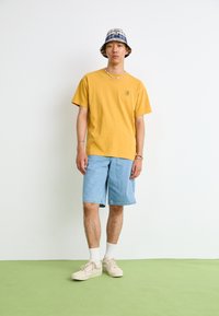 Yellow cotton t-shirt, blue plaid bucket hat, light blue denim shorts, white socks, cream sneakers. Minimalist casual outfit on a green floor.