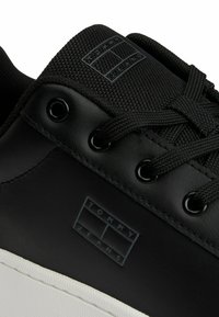 Zapatilla negra con parte superior de cuero, lengüeta de tela negra texturizada y suela de goma blanca. Incluye un detalle de logotipo en relieve sutil.