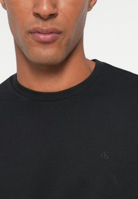 Bărbat cu piele maro medie care poartă un tricou negru cu decolteu rotund și un mic logo „cK” în relief pe piept.