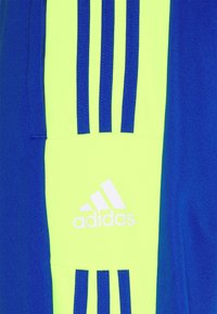 Gul tygpanel med blå vertikala ränder och vit Adidas-logotyp, omgiven av blått tyg på båda sidor.