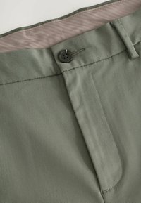 Grüne Hose aus strukturiertem Stoff, mit einem Knopfverschluss und einem glatten, hellen Taillenbund. Saubere Linien und taillierte Passform.