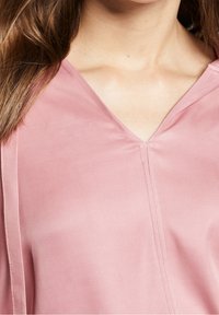 Rosa Bluse mit V-Ausschnitt. Glattes Material, das ein einfaches Design mit klaren Linien und feinen Nähten entlang des Ausschnitts aufweist.