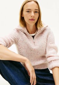 Femme jeune avec les cheveux blonds courts portant un pull tricoté lilas à motifs avec une fermeture éclair à l'avant et un jean bleu foncé, assise sur un fond blanc.