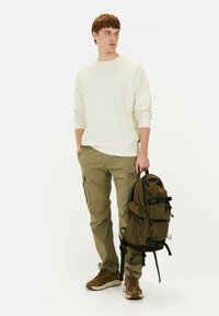 Leichtes Creme-Sweatshirt, olivgrüne Cargo-Hose, braune Sneakers. Hält einen dunkelgrünen Rucksack mit mehreren Taschen und Riemen.