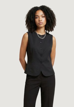 Donna con capelli ricci che indossa un gilet nero senza maniche con bottoni, pantaloni neri e una collana di catena d'argento su uno sfondo semplice.