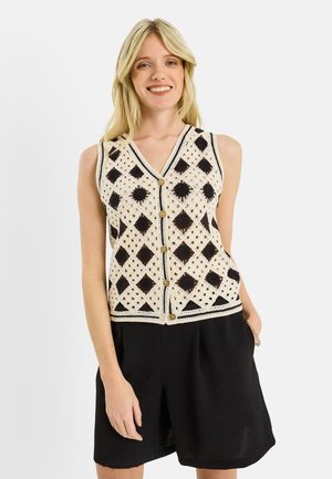 Mujer sonriente que lleva un chaleco de punto sin mangas con un patrón de rombos en negro y beige y botones de madera, combinado con pantalones cortos negros y sueltos.