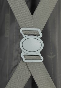 Šedé textilní popruhy se protínají s plastovou šedou sponou, připevněnou na decentním tmavém pozadí s jemnými vzory.