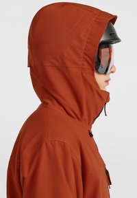 Oranje hoodie-jas met een gestructureerde afwerking, voorzien van een zijritssluiting en een verstelbaar trekkoord. Draagt ski-goggles ter bescherming.