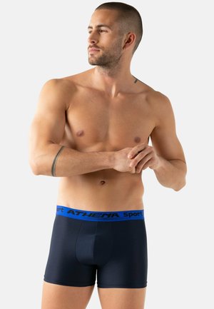 Homme torse nu avec des tatouages, portant un boxer noir avec une ceinture bleue portant l'inscription « ATHENA Sport », joignant les mains et regardant vers la gauche.
