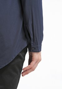 Mørkeblå langærmet skjorte med en glat tekstur, knapmanchetter og en sideslidsdesign. Båret med mørke bukser, der viser skjortens detaljer.