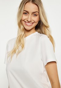Bílý top s krátkým rukávem vyrobený z hladkého materiálu s kulatým výstřihem. Obsahuje jednoduché švy bez vzorů nebo ozdob.