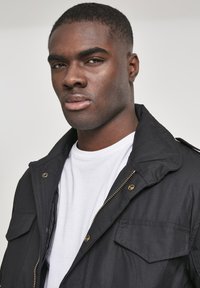Black jacket with a zip-up front, metal hardware, and two chest pockets, worn over a white T-shirt. Smooth fabric and structured design.