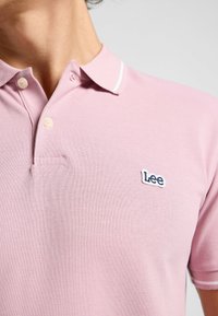 Chemise polo rose en tissu texturé, avec un accent de col blanc, deux boutons et un petit logo brodé Lee sur le côté gauche.