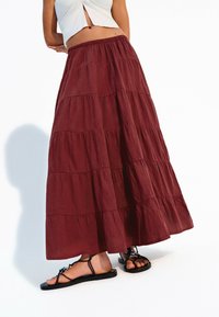 Burgundy, lager maxikjol med elastisk midjeband och luftig textur, tillsammans med svarta, platta sandaler. Har subtila horisontella sömmar.