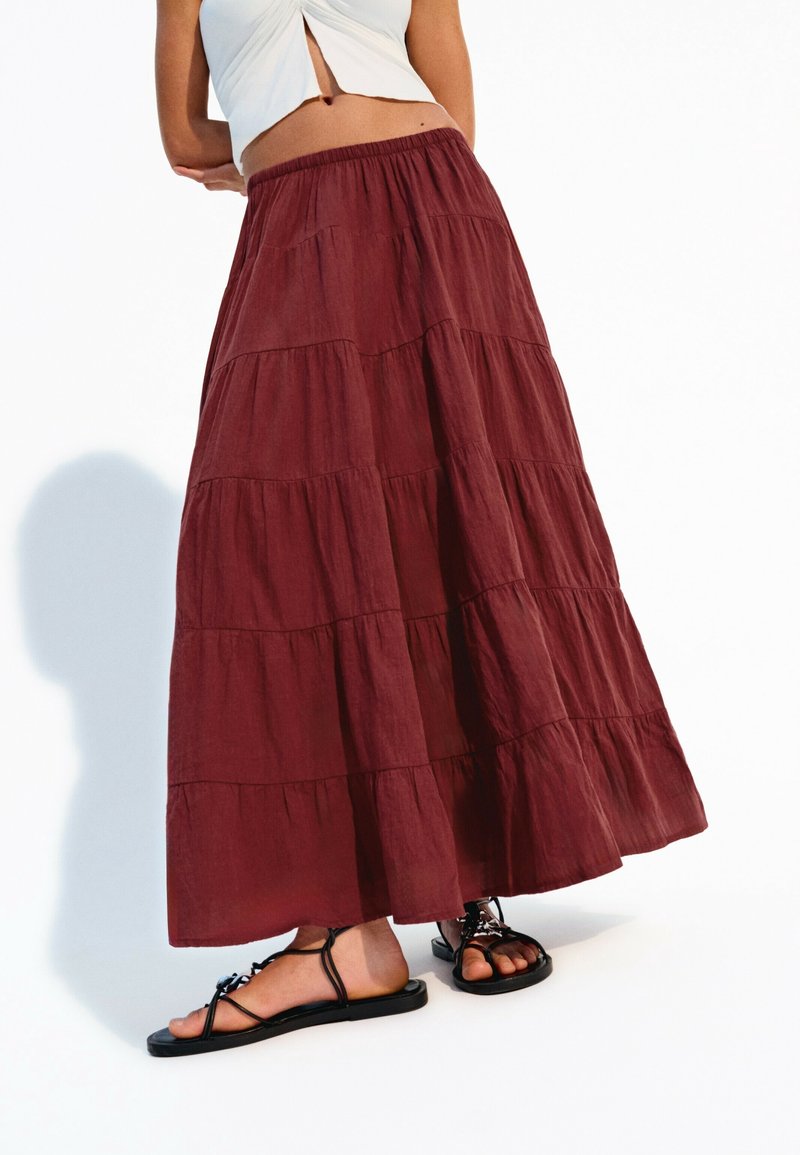 Burgundy, lager maxikjol med elastisk midjeband och luftig textur, tillsammans med svarta, platta sandaler. Har subtila horisontella sömmar.