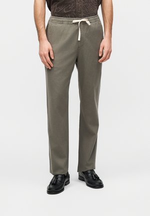 Férfi, aki olívazöld megkötős nadrágot visel, fekete bojtosszőrbakancsokkal, egy egyszínű világos háttér előtt áll.