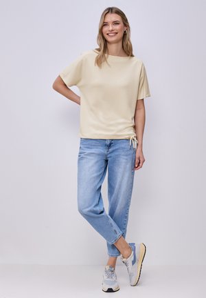 Femme souriante portant un haut beige à manches courtes, un jean bleu clair et des baskets grises, debout avec une jambe croisée et la main sur la hanche.
