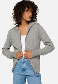 Veste à capuche zippée grise avec une texture douce, dotée d'une capuche et de poignets côtelés, associée à un jean bleu clair.