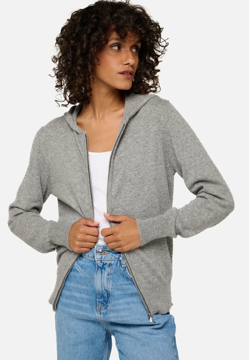 Veste à capuche zippée grise avec une texture douce, dotée d'une capuche et de poignets côtelés, associée à un jean bleu clair.