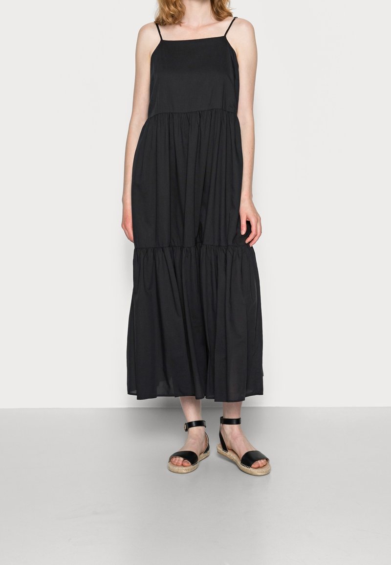 Robe maxi noire sans manches avec un corsage ajusté et une jupe évasée à volants. Porte des sandales noires avec des brides à la cheville et des semelles tissées.