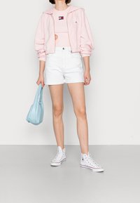 Roze fleece hoodie met rits, lichtroze mouwloos topje, witte denim shorts, blauwe tas en witte hoge sneakers met accenten.