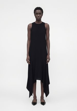 Femme debout, portant une robe noire sans manches à ourlet asymétrique et des chaussures noires pointues, sur un fond blanc uni.