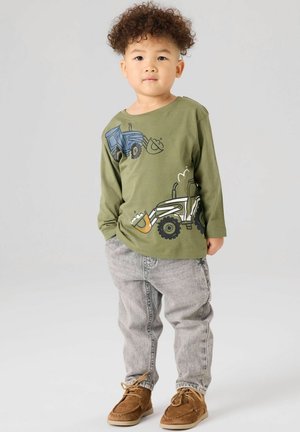 Toddler with curly hair wearing green long-sleeve shirt with tractors, gray jeans, and brown shoes, standing against plain light background.