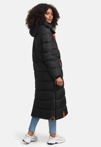 Black long puffer coat with a hood, quilted design, featuring orange accents and zippers. Model wears light blue jeans and white sneakers.