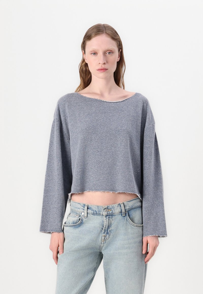 Blusa cinza de manga longa cortada, com um decote largo e detalhes de bainha crua, combinada com jeans de lavagem clara. Textura suave, design casual.