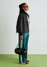 Black, button-up jacket with a fur-lined collar, paired with teal, velvet joggers featuring three white stripes. Black handbag and dark sneakers.