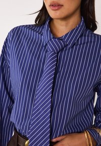 Camicia a righe blu e bianche con colletto e dettaglio del nodo al collo, realizzata in un tessuto morbido. Il modello indossa bracciali dorati.