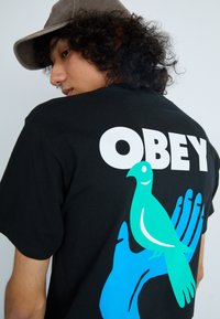Must värvi T-särk, millel on suur valge tekst "OBEY" ja sinine käsi koos rohelise linnuga, sile tekstuur, lühikesed varrukad.