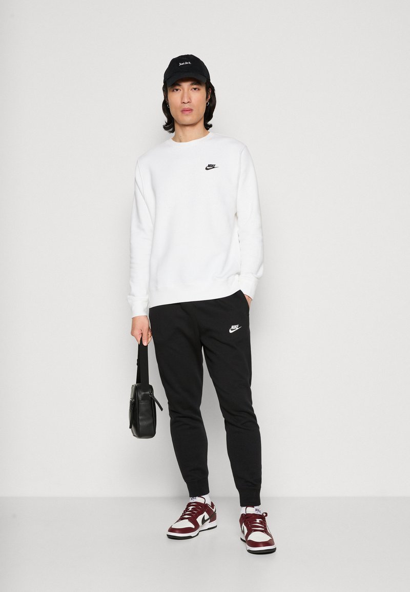 Sudadera blanca con el logo de Nike en negro, pantalones de chándal negros con el logo de Nike, gorra negra y zapatillas burdeos con detalles en blanco. Sosteniendo una bolsa negra.