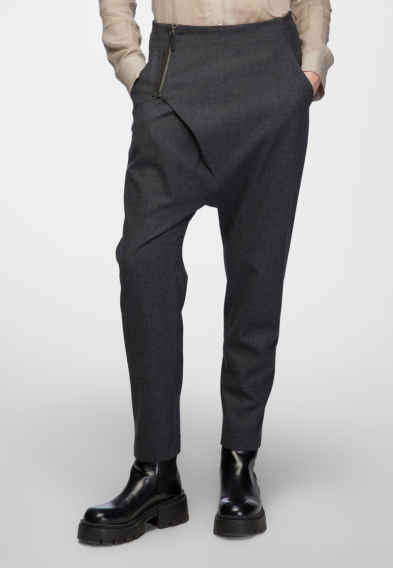 Dark grey, tailored trousers with a unique draped design, zippered front pocket, and tapered leg. Worn with black chunky ankle boots.