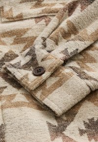 Veste en laine avec une base beige ornée de motifs géométriques marron et rouille, un poignet replié et un accent de bouton noir sur la manche.