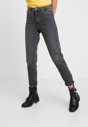 Person trägt hochgekrempelte dunkelgraue Jeans, schwarze Lederstiefel mit Schnalle und ein gelbes, geripptes ärmelloses Oberteil, steht mit gespreizten Beinen.