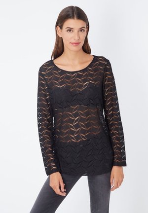 Top negro de encaje de mangas largas con un patrón geométrico. Cuello redondo y ajuste holgado, combinado con pantalones grises.
