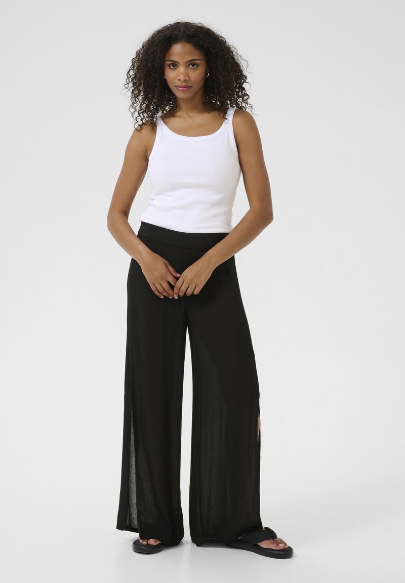 Pantalons noirs à jambe large avec des fentes sur les côtés et une texture fluide, associés à un débardeur blanc ajusté. Le modèle se tient devant un fond uni.