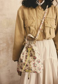 Blomstret bucket taske med en beige bund, gul snøre og gyldent hardware. Båret sammen med lagdelte tøj med lommer.