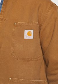 Hnědá plátěná bunda s náprsní kapsou a bílou nálepkou a oranžovým logem, zpevněnými švy a detaily zlaté barvy na knoflících.