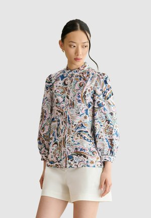 Mujer con cabello negro recogido lleva una blusa de manga larga con estampado floral y pantalones cortos blancos, de pie frente a un fondo liso y claro.