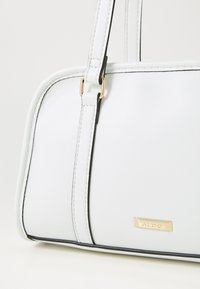 Sac à main en simili cuir blanc avec plaque logo dorée, forme structurée, souligné de piping noir, doté de doubles anses et de détails de couture.