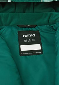 Interno di una giacca verde con un'etichetta nera che riporta "relma", taglia 92. Presenta icone che indicano resistenza all'acqua e calore. Texture liscia.